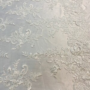 May include: White floral lace fabric with a delicate pattern. The lace is embroidered with small flowers and leaves.