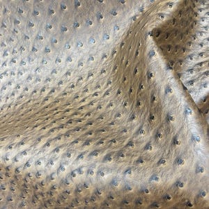 May include: Close-up of a tan leather material with a dimpled texture. The surface has a pattern of small, evenly spaced indentations, creating a unique visual effect. The leather appears soft and pliable, with subtle variations in color.