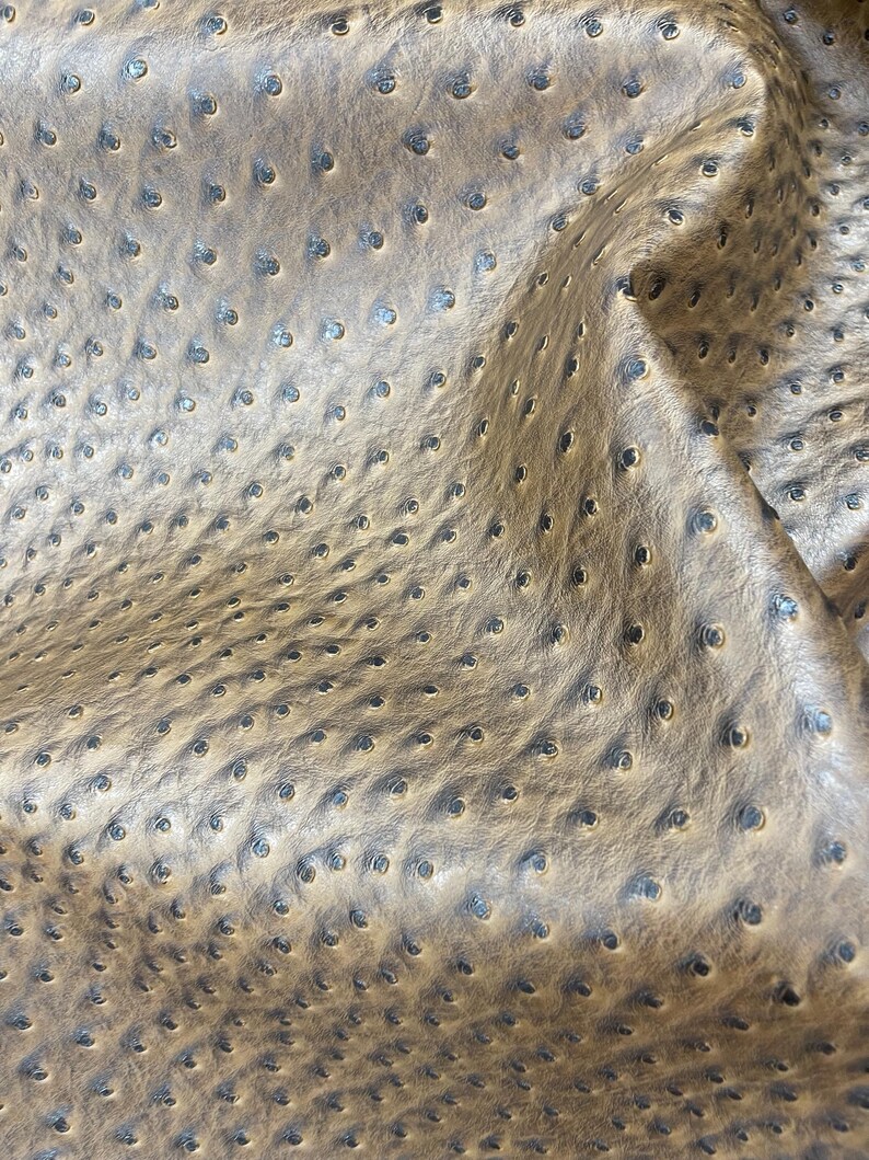 May include: Close-up of a tan leather material with a dimpled texture. The surface has a pattern of small, evenly spaced indentations, creating a unique visual effect. The leather appears soft and pliable, with subtle variations in color.