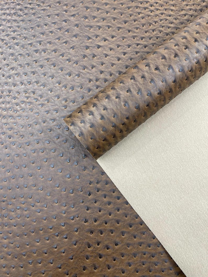 May include: Close-up of brown ostrich leather with a textured surface and small, dark-colored dimples. A portion of the leather is rolled, revealing a beige, smooth backing. The material is suitable for various crafting projects.