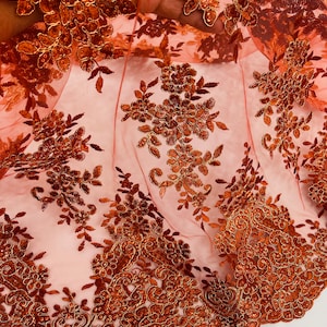 Puede incluir: Tela de encaje floral naranja y dorada con lentejuelas. El encaje tiene un delicado patrón floral con un borde festoneado.