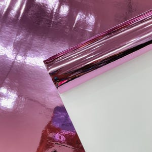 Könnte beinhalten: Eine Rolle glänzendes, pinkfarbenes Metallic-Folienpapier. Die Folie reflektiert das Licht und erzeugt eine glänzende Oberfläche. Ein Abschnitt des Papiers wird abgezogen und gibt eine weiße Unterseite frei. Das Papier wird wahrscheinlich für Bastelarbeiten oder Dekorationszwecke verwendet.