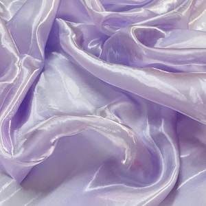 Peut inclure: Gros plan sur un tissu fluide et irisé de couleur lavande douce. Le matériau a un éclat soyeux, avec des plis et des courbes qui captent la lumière, créant des reflets et des ombres. Le tissu semble léger et délicat.