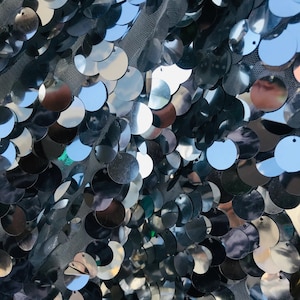 May include: Close-up of a silver sequined fabric with round sequins. The sequins are arranged in a random pattern and create a shimmering effect.