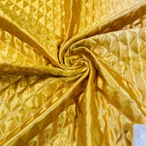 May include: A close-up shot of a golden yellow quilted fabric with a diamond pattern. The fabric is folded and swirled, showcasing its shiny, satin-like texture. The stitching creates a grid of diamond shapes across the material.