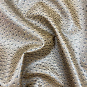 May include: Close-up of a textured, tan-colored fabric with a pattern of small, evenly spaced perforations. The material has a subtle sheen and a slightly wrinkled appearance, suggesting it is flexible and pliable.