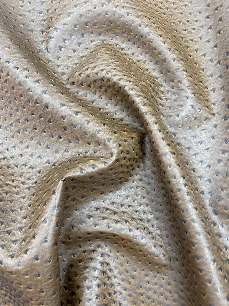 May include: Close-up of a textured, tan-colored fabric with a pattern of small, evenly spaced perforations. The material has a subtle sheen and a slightly wrinkled appearance, suggesting it is flexible and pliable.