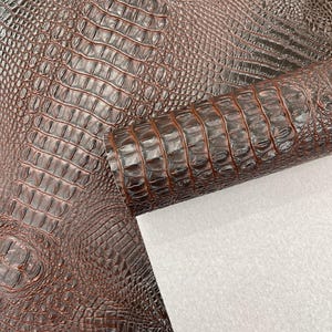 May include: Close-up of brown faux crocodile leather. The material has a textured, scaled pattern and a rolled section revealing a white backing. The color is a rich, dark brown with highlights.