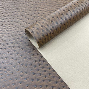 May include: A close-up of a roll of brown faux leather with a textured ostrich pattern. The material is a rich, warm brown with darker accents in the dimples, and a light beige backing is visible.