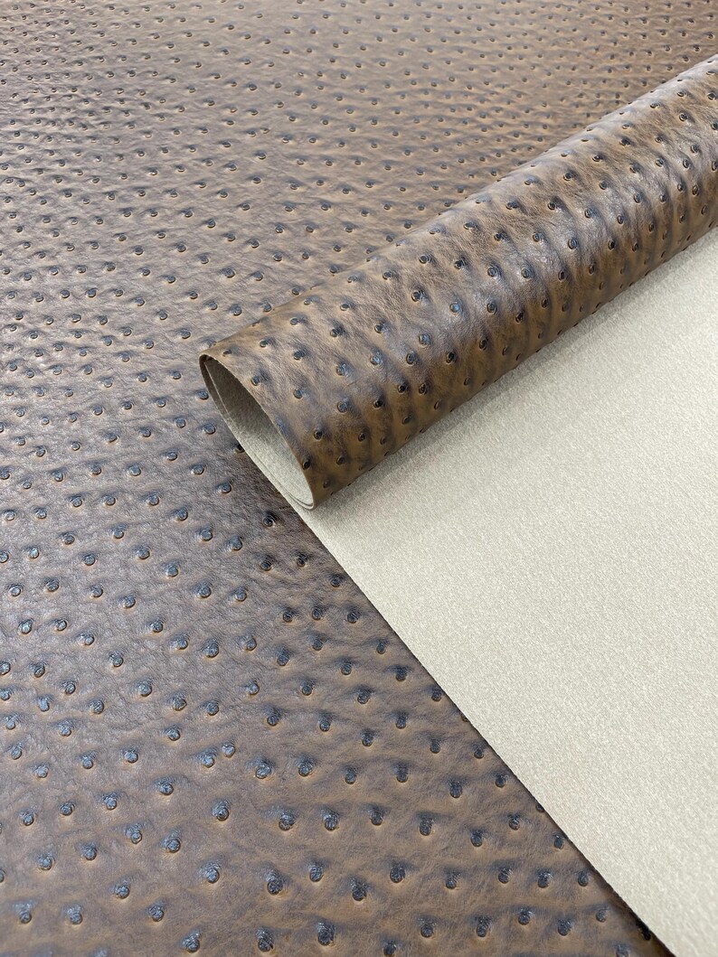 May include: A close-up of a roll of brown faux leather with a textured ostrich pattern. The material is a rich, warm brown with darker accents in the dimples, and a light beige backing is visible.