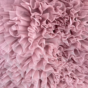 Puede incluir: Primer plano de una tela con volantes en color rosa empolvado. La tela parece ser tul, con múltiples capas que crean un efecto texturizado y voluminoso. Los pliegues y las ondas de la tela crean una estética suave y romántica.