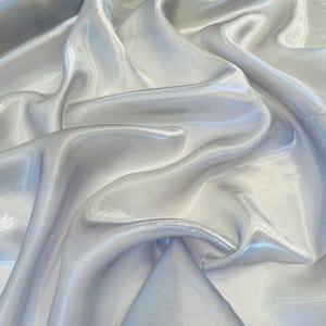 Peut inclure: Gros plan sur un tissu fluide et irisé dans les tons argent et blanc. Le matériau a un éclat brillant, avec des plis et des courbes douces qui captent la lumière, créant des reflets et des ombres. Le tissu semble léger et délicat.