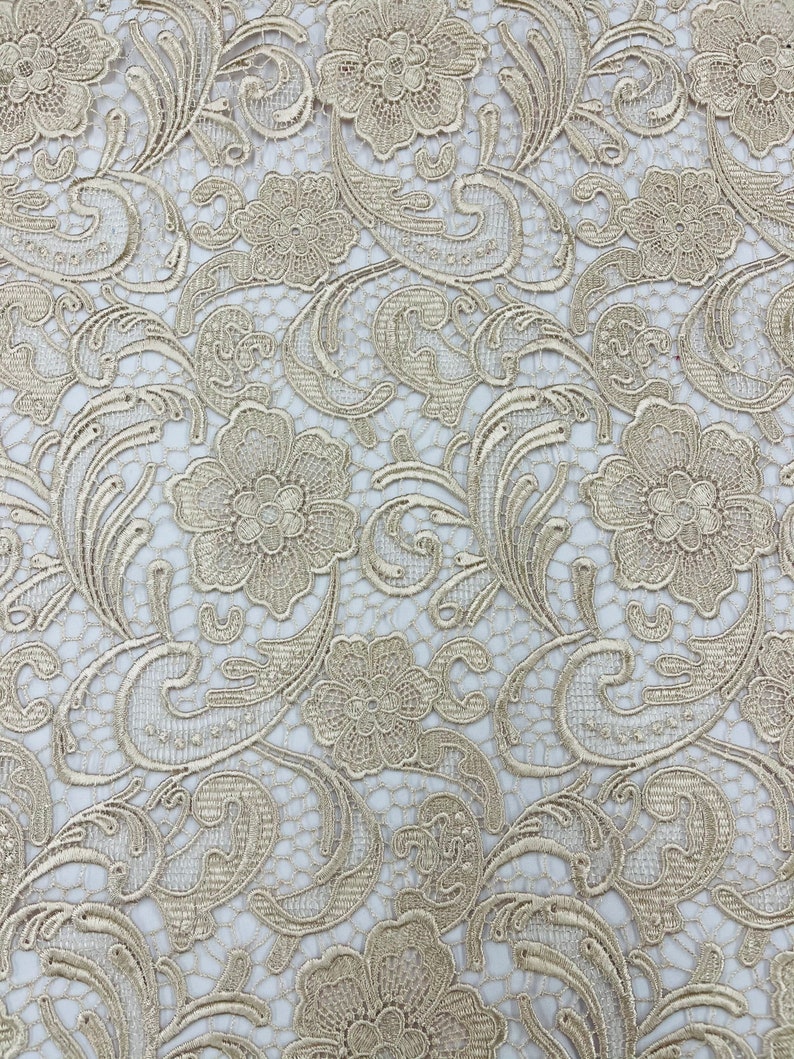 May include: A close-up of a beige lace fabric with a floral pattern. The lace has a delicate, intricate design with small flowers and swirls.