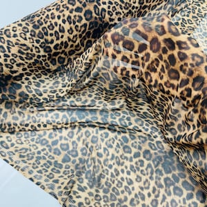 Könnte beinhalten: Eine Rolle transparenten Stoffes mit Leopardenmuster. Der Stoff hat einen beigefarbenen Hintergrund mit dunkelbraunen Flecken, wodurch ein klassisches Tiermuster entsteht. Das Material wirkt leicht und fließend.