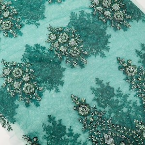 May include: Emerald green lace fabric with floral embroidery. The fabric features clusters of beaded flowers and sequins, creating a textured and decorative design. The lace is semi-transparent, with a light teal background.