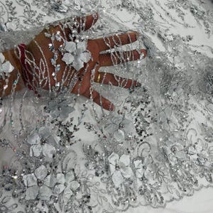 May include: Silver and white floral lace fabric with sequins and 3D flowers. The fabric has a delicate, intricate design and is perfect for adding a touch of elegance to any project.