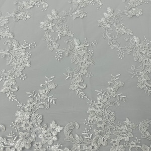 May include: Close-up of white floral lace fabric. The lace features detailed embroidered flowers, leaves, and swirling patterns. The design is delicate and intricate, with a scalloped edge along the bottom. The fabric is on a light grey background.