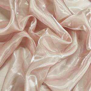 Peut inclure: Gros plan sur un tissu rose clair, fluide et brillant. Le matériau présente un aspect chatoyant, presque irisé, avec des plis doux et des reflets qui captent la lumière. Le tissu semble léger et délicat.