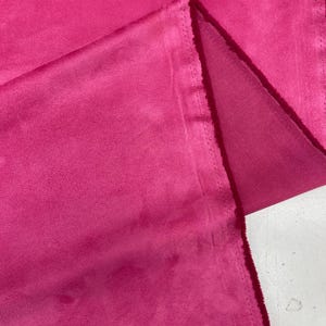 May include: Close-up of a vibrant fuchsia fabric. The material appears to be a soft, suede-like texture, with visible folds and a stitched edge. The colour is consistent throughout the fabric, creating a rich, saturated appearance. The fabric is likely used for apparel or home decor.