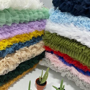 May include: A collection of colorful tulle fabric in various shades, including white, green, purple, yellow, and blue. The fabric is arranged in layered stacks, with small potted cacti in the foreground.