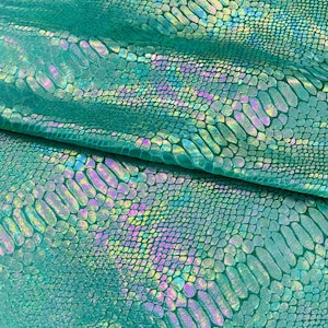 Puede incluir: Primer plano de una tela turquesa con un patrón de piel de serpiente. La tela tiene una calidad brillante e iridiscente, con toques de morado, amarillo y azul. Las escamas son detalladas y texturizadas, creando una apariencia realista. La tela está doblada, mostrando la textura y el brillo.