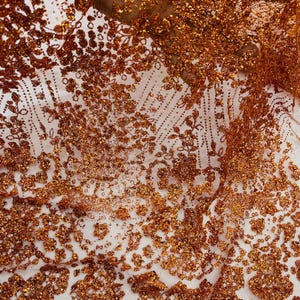 Può includere: Primo piano di un tessuto di pizzo scintillante color rame. Il pizzo presenta intricati motivi floreali ed è pesantemente impreziosito da glitter, creando un effetto strutturato e scintillante. Il tessuto è ideale per l'artigianato e le occasioni speciali.