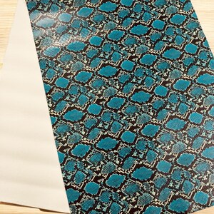 May include: A sheet of faux leather with a teal and black snakeskin pattern. The pattern is made up of diamond shapes with a white outline. A section of the sheet is folded over, revealing a white backing.