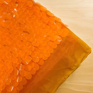 Könnte beinhalten: Orangener Stoff mit einem Muster aus überlappenden, runden Pailletten. Der Stoff ist durchsichtig und hat einen leicht schimmernden Glanz.