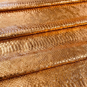 Op de afbeelding: Gouden metallic stof met een slangenhuidpatroon. De stof is in meerdere lagen gevouwen, waardoor de textuur en glans van het materiaal zichtbaar worden.