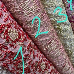 May include: Close-up of several rolls of textured fabric in various colors. The fabrics include red and gold, pink and gold, and pale gold. The fabrics have a crinkled appearance and a metallic sheen, suggesting they are intended for decorative or apparel use.