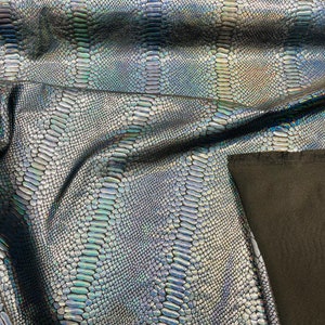 May include: Close-up of iridescent black fabric with a snake skin pattern. The fabric is folded and draped over a black surface.