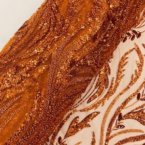 May include: Close-up of a shimmering, orange-gold fabric. The material is heavily embellished with sequins and beads, creating a textured, flowing effect. The design includes a detailed pattern of swirling lines and leaf-like shapes.