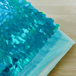 May include: A turquoise blue pillow with a decorative pattern of overlapping sequins. The sequins are arranged in a wave-like pattern.