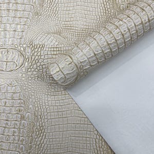 May include: Ivory-coloured alligator-textured leather. The leather is rolled up, revealing a white backing. The surface has a detailed pattern of scales and ridges, creating a realistic crocodile skin appearance. The colour is a soft, neutral tone.