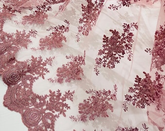 May include: A sheer, light pink fabric with intricate burgundy floral embroidery. The design features detailed flowers and leaves, creating a delicate and textured pattern. The edge of the fabric has a scalloped border with rose-like motifs.
