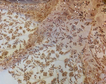 May include: Close-up of a gold and beige beaded fabric with a floral pattern. The fabric is sheer and has a delicate, sparkly texture.
