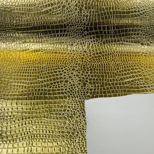 May include: Close-up of a sheet of gold-colored faux leather with a crocodile skin texture. The material has a shiny, metallic finish and a repeating pattern of small, rectangular scales. A white, unfinished edge is visible.