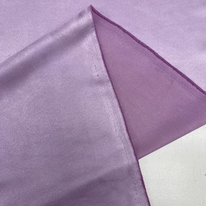 May include: A close-up of a lavender-colored satin fabric. The fabric is folded, revealing a seam and a contrasting lighter shade of lavender on the underside. The satin has a smooth, shiny texture.