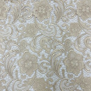 May include: A close-up of a delicate, gold-colored lace fabric with a floral pattern. The lace has a fine mesh background and intricate floral designs.