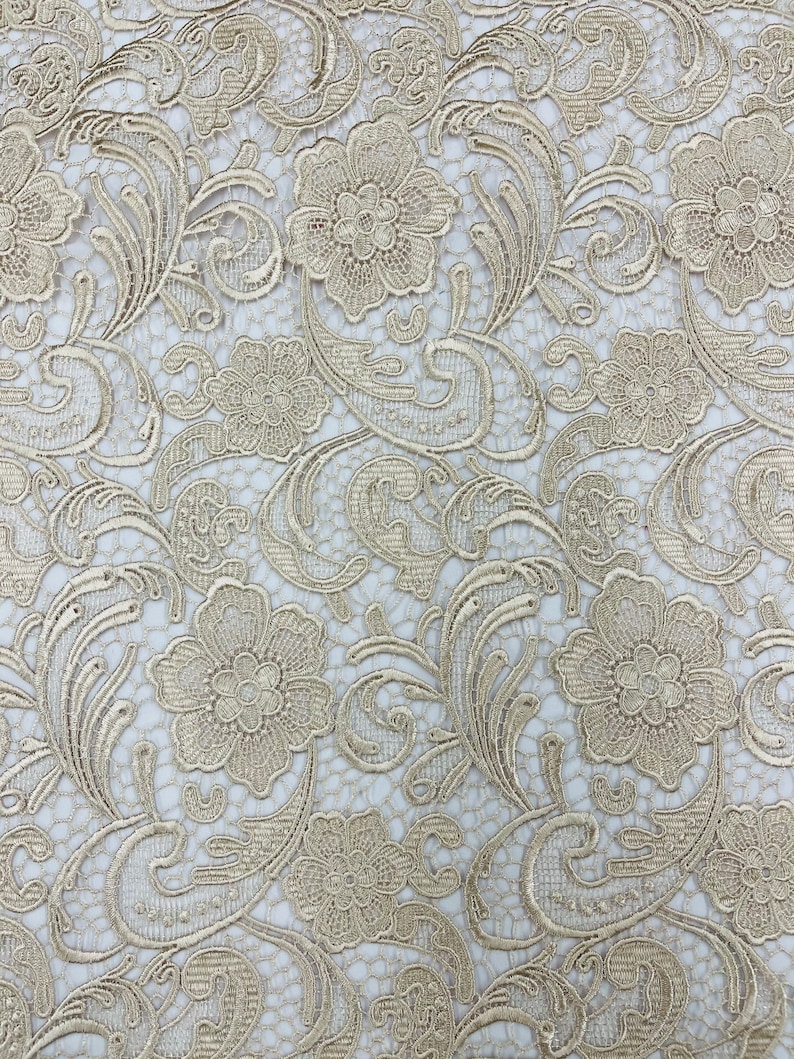 May include: A close-up of a delicate, gold-colored lace fabric with a floral pattern. The lace has a fine mesh background and intricate floral designs.