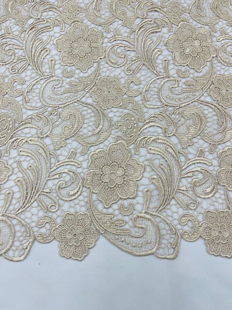 May include: Beige lace fabric with a delicate floral pattern. The lace features intricate swirls and small flowers.