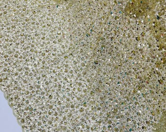 May include: Close-up of a shimmering fabric with a dense pattern of sequins and beads. The fabric has a light gold color with iridescent accents, creating a sparkling effect. The edges are scalloped.