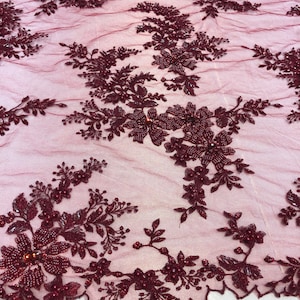 May include: Burgundy beaded lace fabric featuring a floral design. The lace is delicate and intricate, with small beads and sequins throughout. The fabric is ideal for adding a touch of elegance to any project.