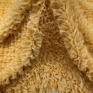 Pode incluir: Close-up de um tecido com textura franzida em um tom amarelo dourado quente. O material parece ser em camadas, criando um efeito denso e volumoso. As dobras e ondas do tecido capturam a luz, destacando sua natureza delicada.