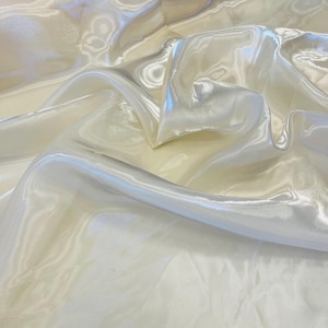 Puede incluir: Primer plano de una tela crema brillante con una textura sedosa. La tela está drapeada en pliegues suaves, creando luces y sombras. El material tiene un brillo sutil, lo que sugiere que es un material ligero.