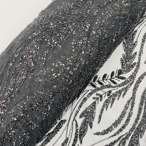 May include: Two rolls of fabric are shown. One is black, densely covered in sequins and beads, giving a textured, sparkling appearance. The other is white with a black, leaf-like design. These fabrics are likely intended for clothing or decorative purposes.
