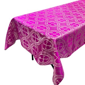 May include: A pink tablecloth with a white cross pattern. The tablecloth is made of a brocade fabric and has a shiny finish.