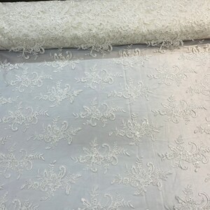 May include: White floral lace fabric with a delicate pattern. The lace is soft and sheer, with a subtle shimmer. It is perfect for adding a touch of elegance to any project.