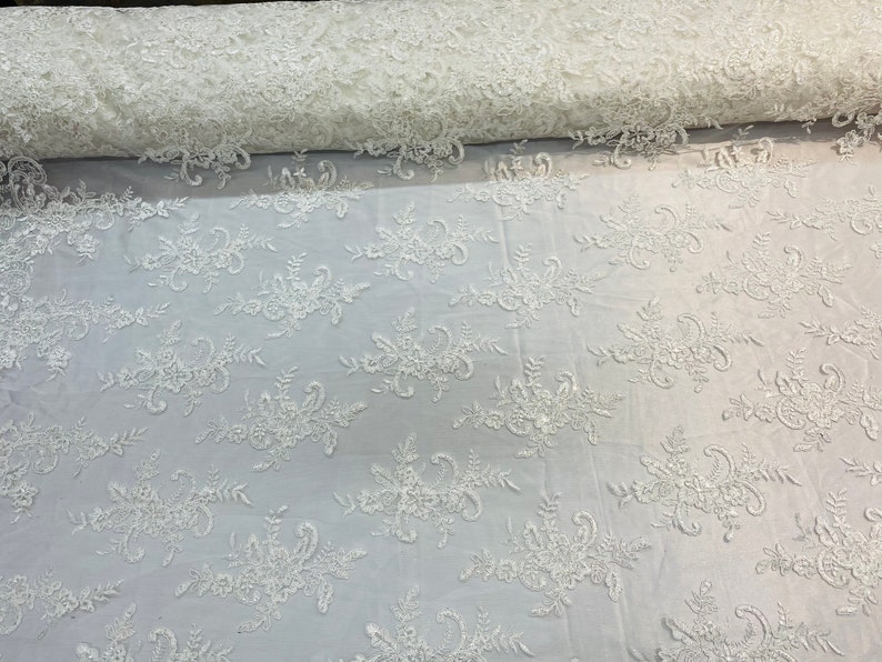 May include: White floral lace fabric with a delicate pattern. The lace is soft and sheer, with a subtle shimmer. It is perfect for adding a touch of elegance to any project.
