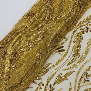 May include: Close-up of gold sequined fabric with a flowing, textured design. The fabric has a shimmering, metallic appearance, with intricate patterns of sequins and beads. The background is white, highlighting the gold embellishments. The fabric is likely used for apparel or decorative purposes.